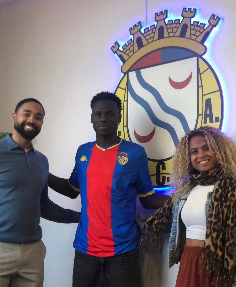 A player in a blue and red FC Alverca shirt standing between a man and a woman in front of the club crest