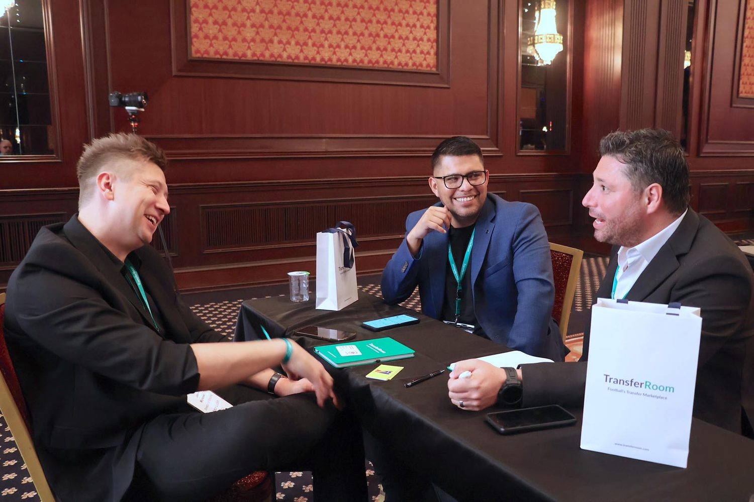 Three men talking and laughing around a meeting table at a TransferRoom event