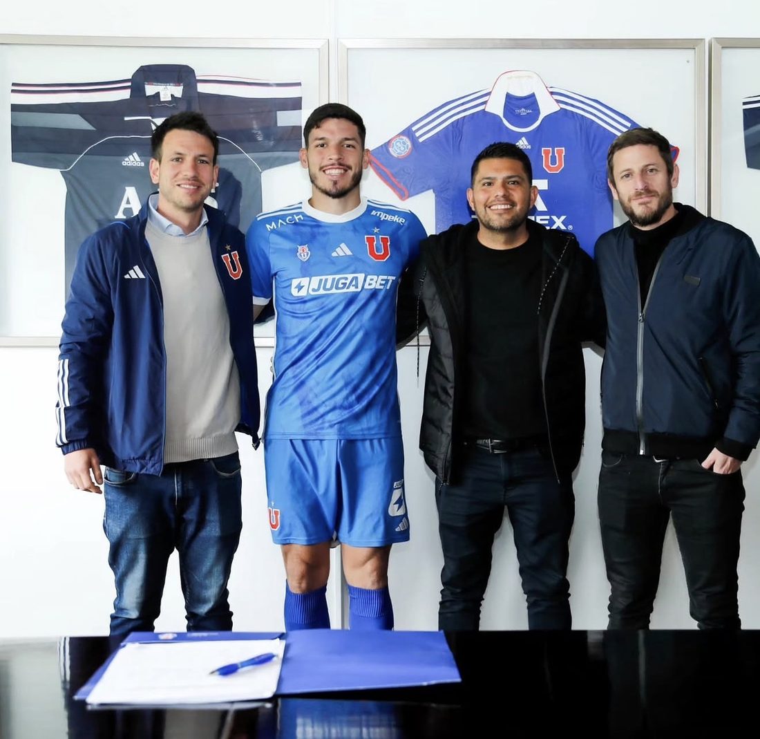 A player in a blue Universidad de Chile kit standing with three men behind a table with a contract and pen